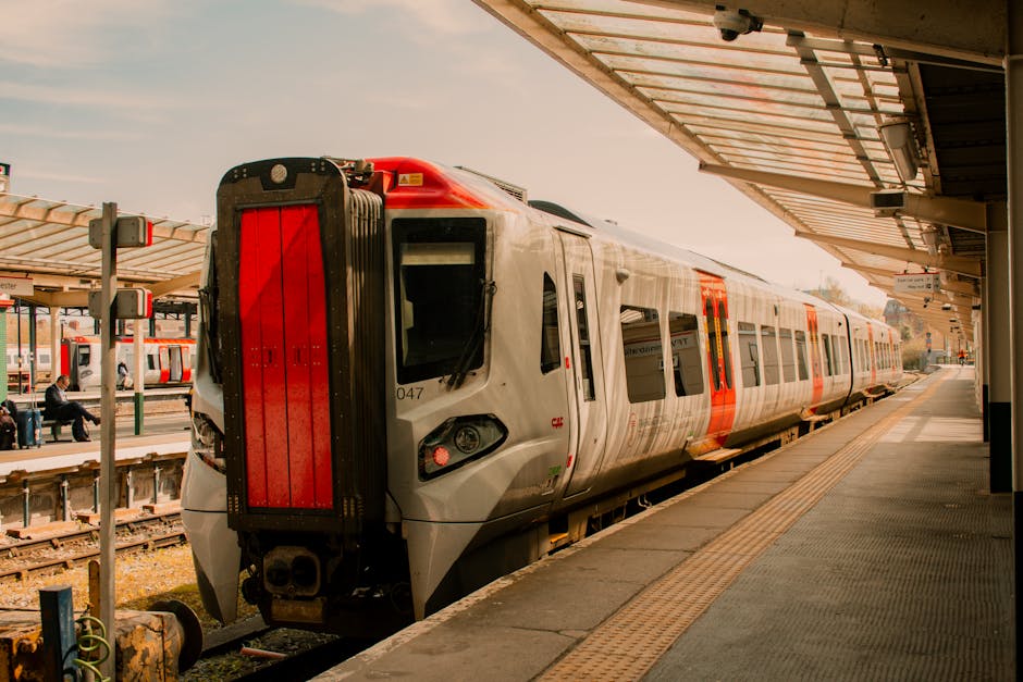 A modern, silver commuter train with orange safety markings is stationary at a platform part of Forest Gate station, which features a covered waiting area with a beige roof supported by metal beams. T