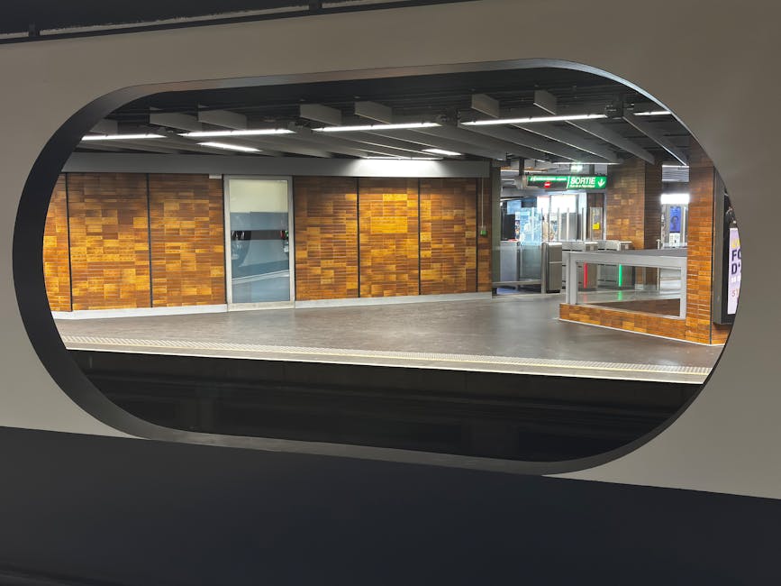 A modern, silver commuter train with orange safety markings is stationary at a platform part of Forest Gate station, which features a covered waiting area with a beige roof supported by metal beams. T